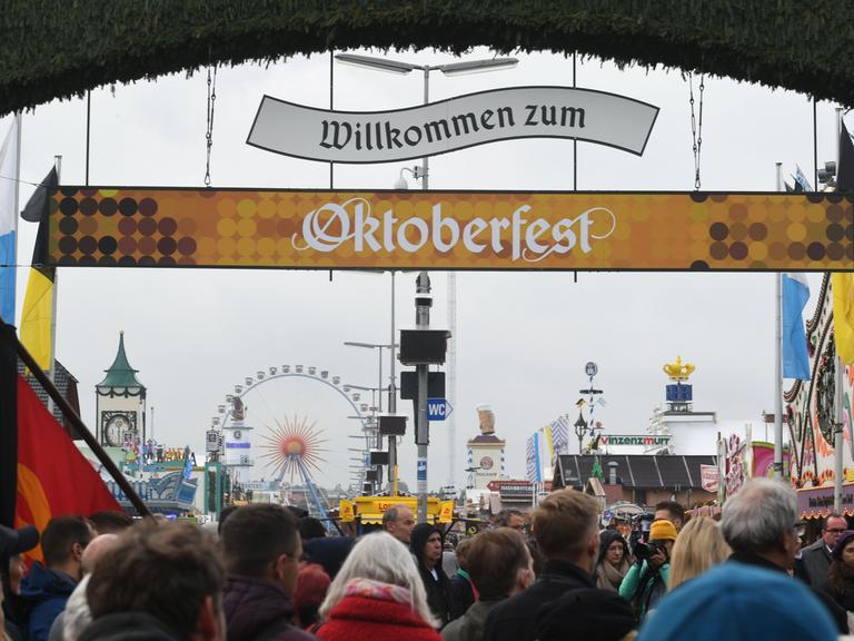 Über den hereinströmenden Besuchern hängen zwei Banner mit der Aufschrift "Willkommen zum Oktoberfest". Im Hintergrund ein Riesenrad und weitere Schausteller-Geschäfte.