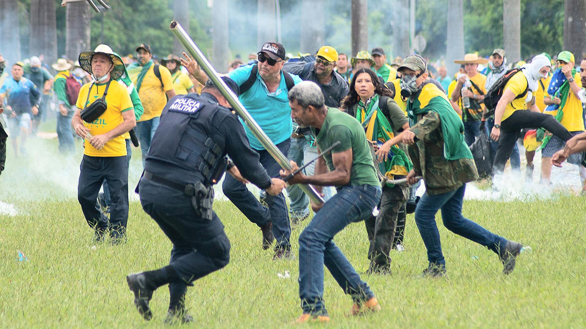 Ein Mann schlägt einen Polizisten mit einer Stange. Dahinter stürmen viele weitere Anhänger vom früheren Präsidenten Bolsonaro heran. Ein Mann schlägt einen Polizisten mit einer Stange. Dahinter stürmen viele weitere Anhänger vom früheren Präsidenten Bolsonaro heran.