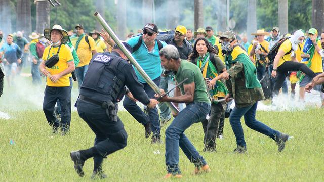 Ein Mann schlägt einen Polizisten mit einer Stange. Dahinter stürmen viele weitere Bolsonaro-Anhänger heran. 