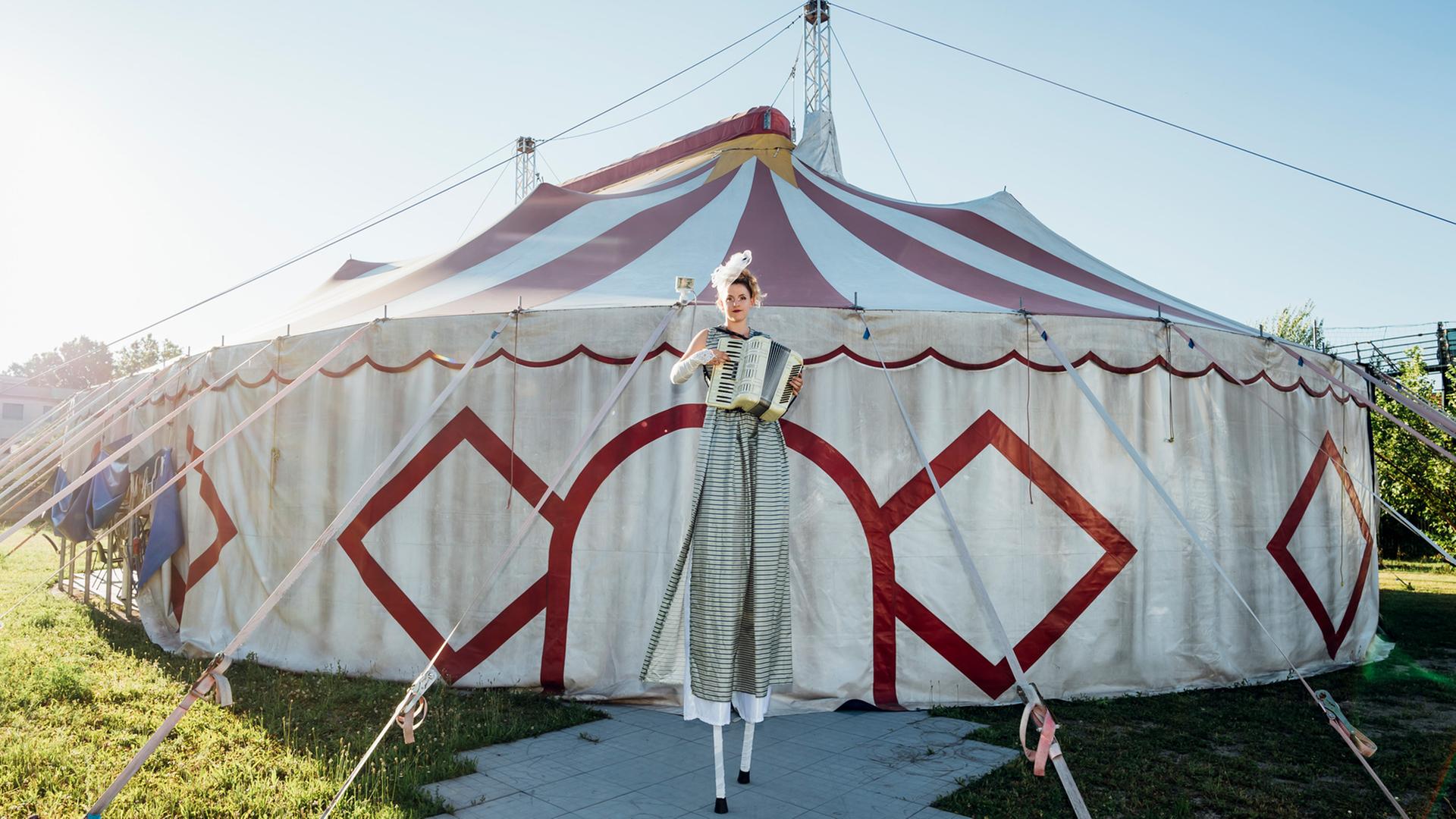 Zirkus bezaubert und ist atemberaubendes Spiel. Wer sind die Menschen, die hier arbeiten? Zu sehen: Eine Frau auf Stelzen steht vor einem Zirkuszelt und spielt Akkordeon. Zirkus bezaubert und ist atemberaubendes Spiel. Wer sind die Menschen, die hier arbeiten? Zu sehen: Eine Frau auf Stelzen steht vor einem Zirkuszelt und spielt Akkordeon.
