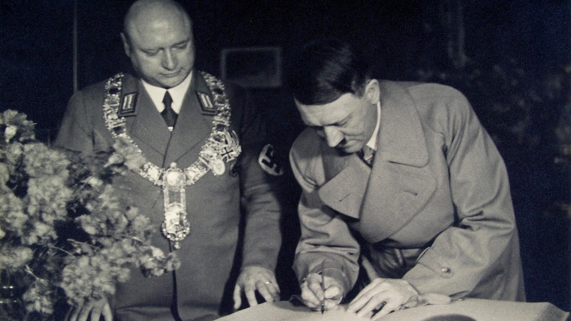 Adolf Hitler schreibt in ein großes Buch, neben ihm steht ein Mann mit Glatze, goldener Bürgermeisterkette und Hakenkreuzbinde. Adolf Hitler schreibt in ein großes Buch, neben ihm steht ein Mann mit Glatze, goldener Bürgermeisterkette und Hakenkreuzbinde.