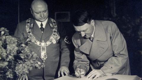 Adolf Hitler schreibt in ein großes Buch, neben ihm steht ein Mann mit Glatze, goldener Bürgermeisterkette und Hakenkreuzbinde.