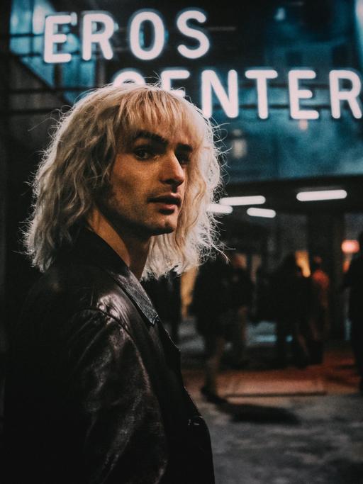 Ein Szenenfoto aus der Amazon-Serie "Luden - Die Könige der Reeperbahn". Zu sehen ist ein junger Mann mit langen blonden Haaren. Hinter ihm ist in der Dunkelheit der Neonschriftzug "Eros Center" zu sehen. Es ist der Schauspieler Aaron Hilmer in der Rolle des Zuhälters Klaus Barkowsky.