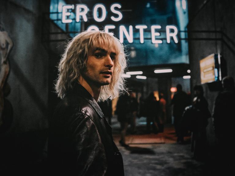 Ein Szenenfoto aus der Amazon-Serie "Luden - Die Könige der Reeperbahn". Zu sehen ist ein junger Mann mit langen blonden Haaren. Hinter ihm ist in der Dunkelheit der Neonschriftzug "Eros Center" zu sehen. Es ist der Schauspieler Aaron Hilmer in der Rolle des Zuhälters Klaus Barkowsky.