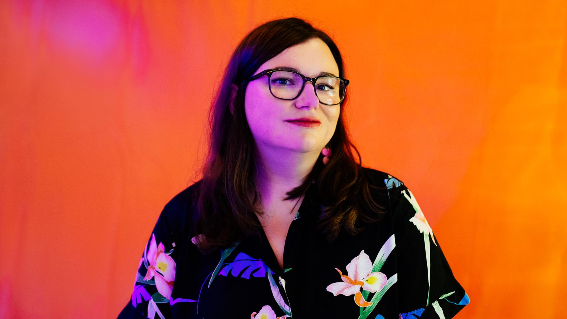 Porträt der Satirikerin Julia Mateus: Sie steht in einer Bluse mit Blumenmotiven vor einem leuchtend orangen Hintergrund. Porträt der Satirikerin Julia Mateus: Sie steht in einer Bluse mit Blumenmotiven vor einem leuchtend orangen Hintergrund.