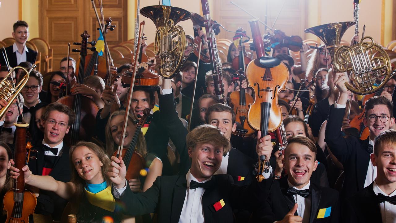 Junge Musikerinnen und Musiker mit ukrainischen und deutschen Flaggen an der Kleidung und an ihren Instrumenten heben in großer Runde ihre Musikinstrumente lachend in die Höhe.
