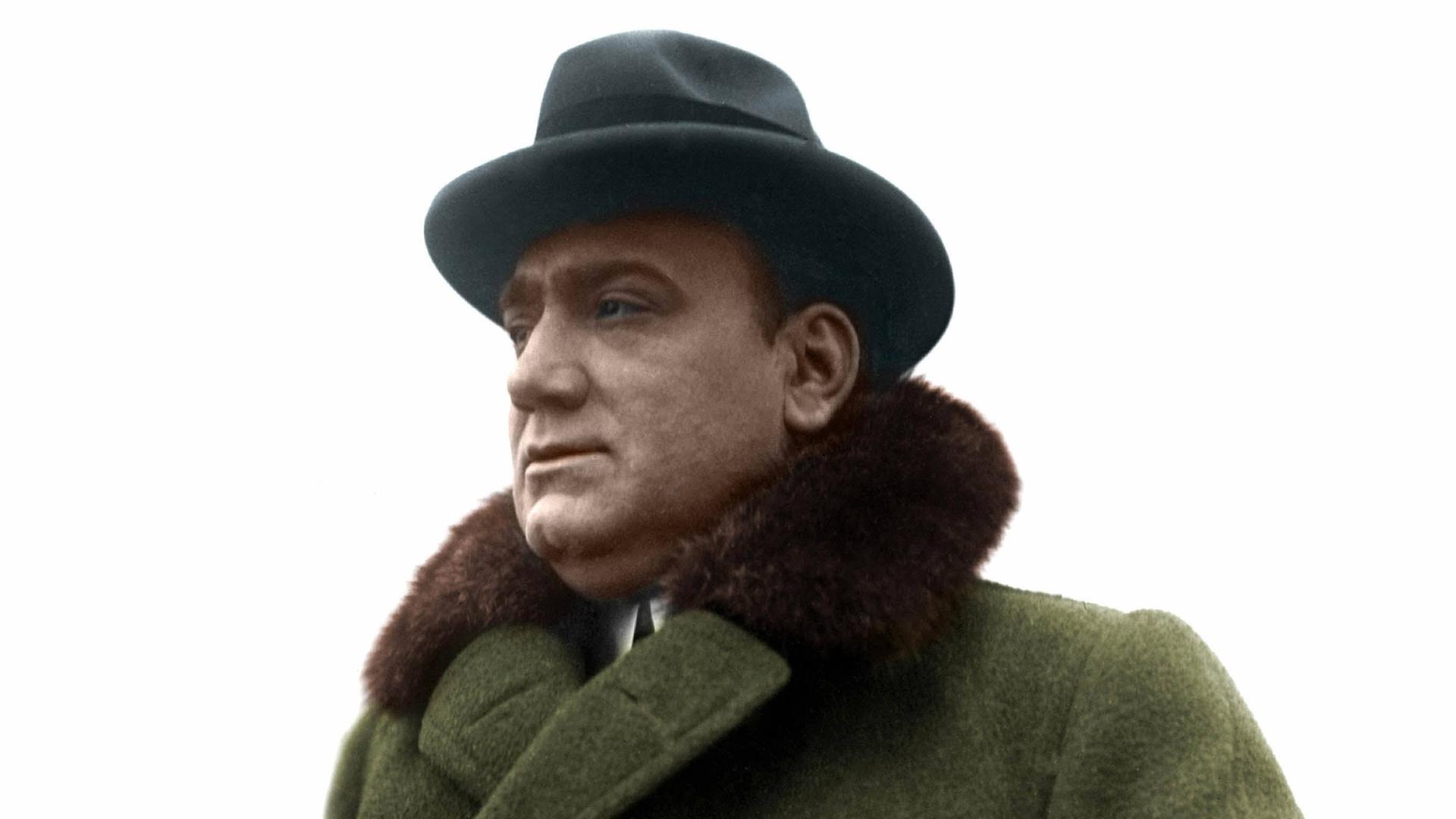 Nachkolorierte Porträtfotografie des Tenors Enrico Caruso (1873-1921) im grünen Mantel mit Pelzbesatz und dunklem Hut.