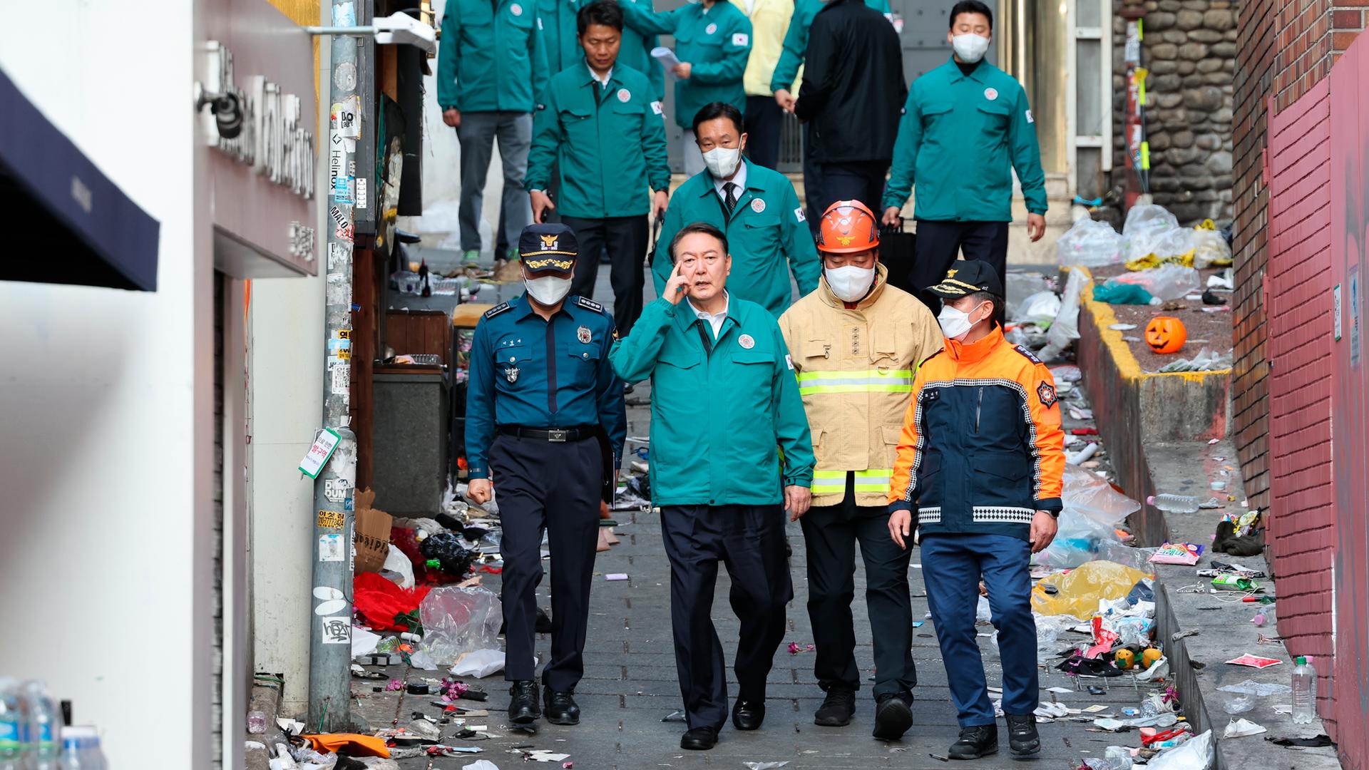 Mehrere Männer, darunter ein Polizist und ein Feuerwehrmann, gehen durch eine enge Gasse, an deren Rändern Abfall liegt. Sie begleiten den südkoreanischen Präsidenten bei dessen Besuch am Unglücksort.