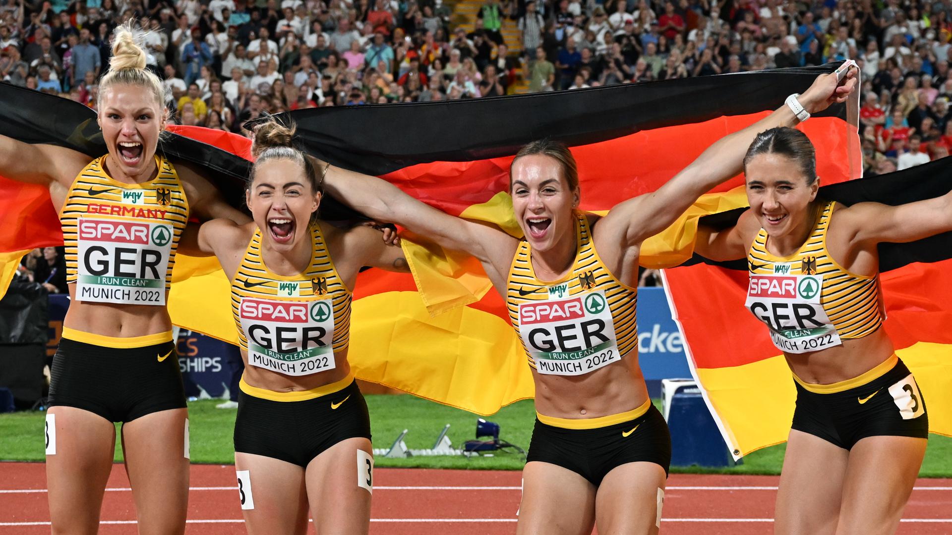 Lisa Mayer, Gina Lückenkemper, Alexandra Burghardt und Rebekka Haase haben eine schwarz-ro-goldene Flagge hinter sich aufgespannt und lachen in die Kamera. Lisa Mayer, Gina Lückenkemper, Alexandra Burghardt und Rebekka Haase haben eine schwarz-ro-goldene Flagge hinter sich aufgespannt und lachen in die Kamera.