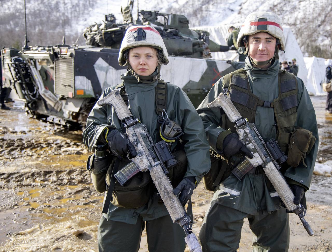 Eine bewaffnete Frau und ein Mann  in Militärkleidung und mit Helm stehen im Schlamm vor einem Panzer.