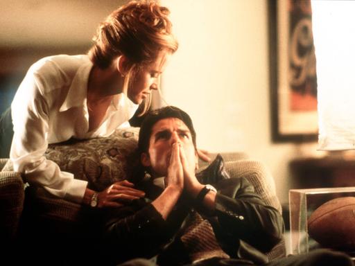 Tom Cruise sitzt mit den Händen vor dem Mund auf einem Sofa, Renée Zellweger beugt sich von hinten über ihn.