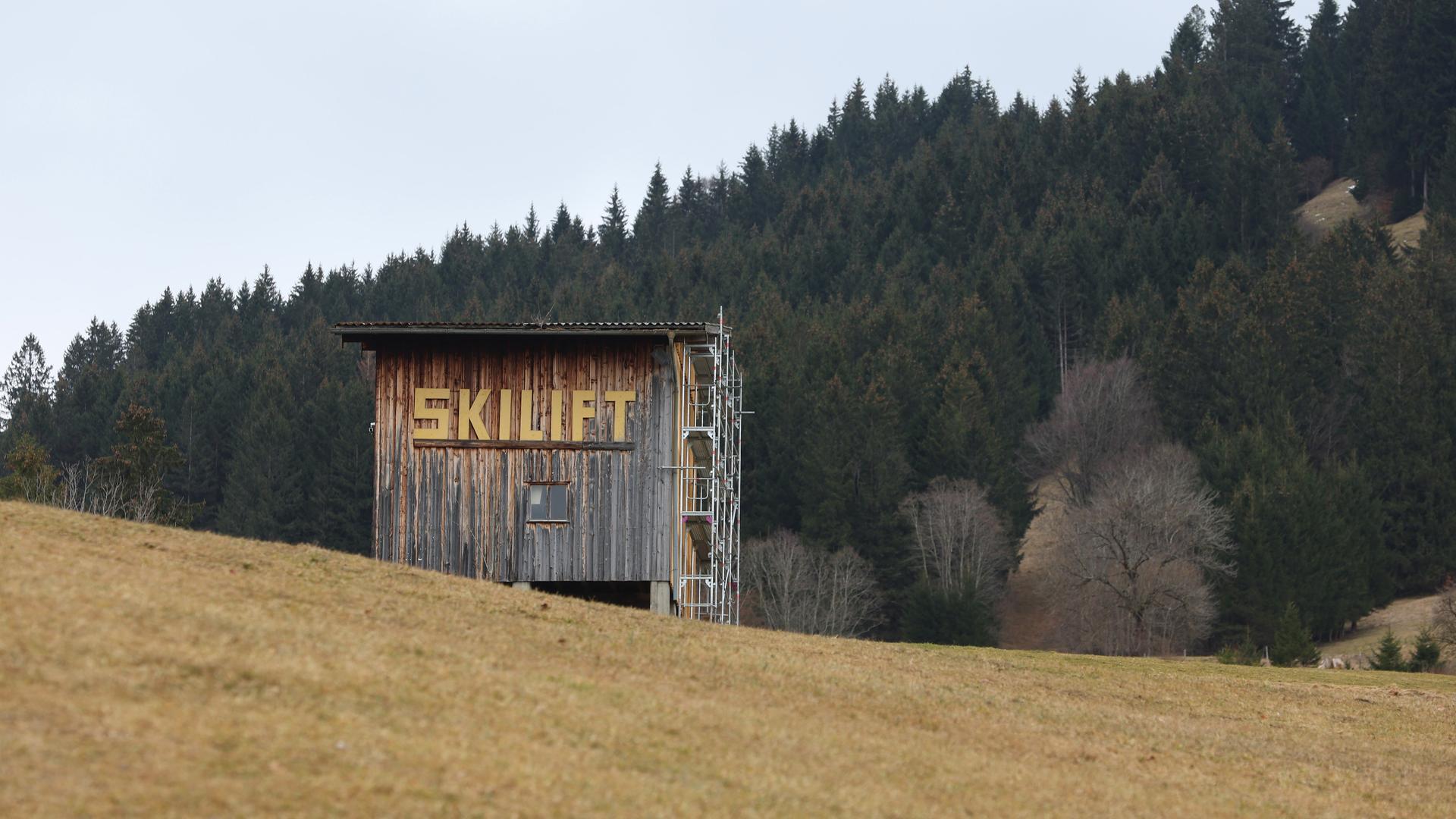 C'è una casa di legno con un'iscrizione su una collina "Sciovia".  La montagna è nuda, senza neve.  Una foresta è visibile sullo sfondo.