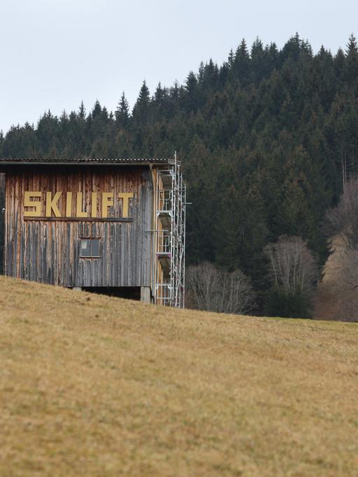 Auf einem Hügel steht ein Holzhaus mit der Aufschrift "Skilift". Der Hügel ist kahl, es liegt kein Schnee. Im Hintergrund sieht man einen Wald.