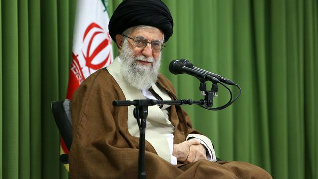 Ali Chamenei sitzt auf einer Bühne. Hinter ihm steht eine iranische Flagge.