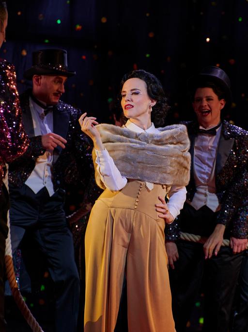 Szenenbild der Jazz-Operette "Die lustige Witwe": Die Schauspielerin Rebecca Nelsen posiert in ihrer Rolle als Hanna Glawari in luxuriös anmutender Kleidung vor dem Ensemble. 
