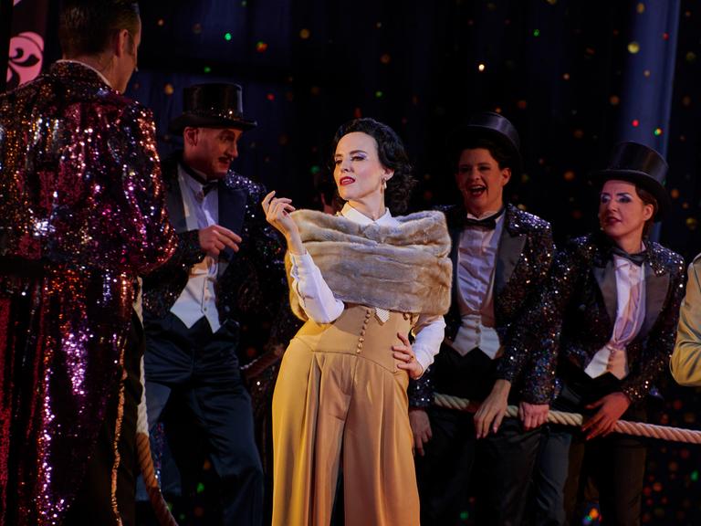 Szenenbild der Jazz-Operette "Die lustige Witwe": Die Schauspielerin Rebecca Nelsen posiert in ihrer Rolle als Hanna Glawari in luxuriös anmutender Kleidung vor dem Ensemble. 