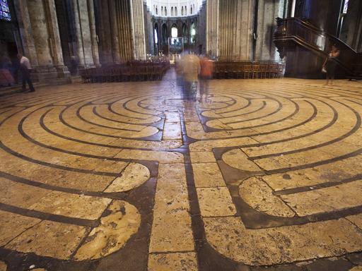 Das Foto zeigt das im Steinboden der Kathedrale von Chartres zu sehende Labyrinth. Im Hintergrund der Altar, unscharf sind zwei Menschen auf dem Labyrinth zu erkennen, die dort gehen.