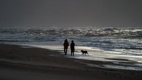 Ein Mann und eine Frau gehen mit einem Hund am Strand von Westerland entlang, an dem sich durch starke Sturmböen das Wasser zu hohen Wellen auftürmt.
