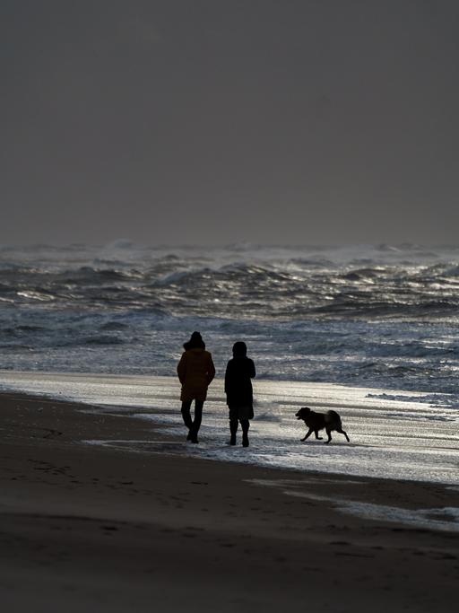 Ein Mann und eine Frau gehen mit einem Hund am Strand von Westerland entlang, an dem sich durch starke Sturmböen das Wasser zu hohen Wellen auftürmt.