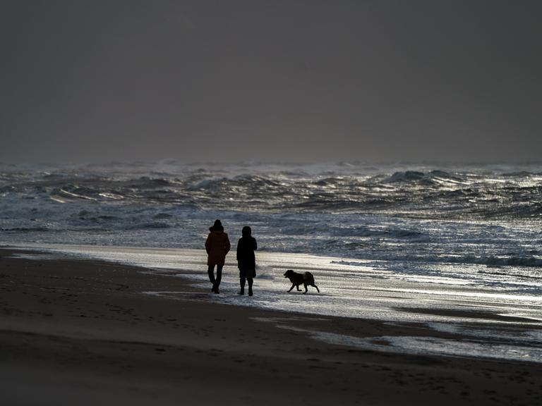 Ein Mann und eine Frau gehen mit einem Hund am Strand von Westerland entlang, an dem sich durch starke Sturmböen das Wasser zu hohen Wellen auftürmt.