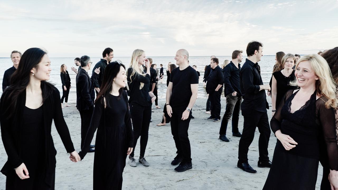 Eine große Gruppe schwarz gekleideter Frauen und Männer stehen in vergnügter Laune verstreut an einem Strand zusammen. Eine große Gruppe schwarz gekleideter Frauen und Männer stehen in vergnügter Laune verstreut an einem Strand zusammen.