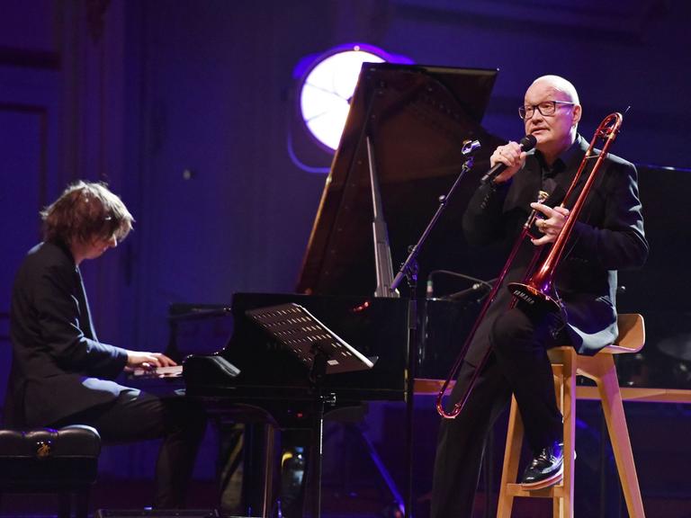 Michael Wollny begleitet Nils Landgren (Posaune) am Klavier auf einer rot-blau ausgeleuchteten Bühne in Hamburg im Jahr 2019. 