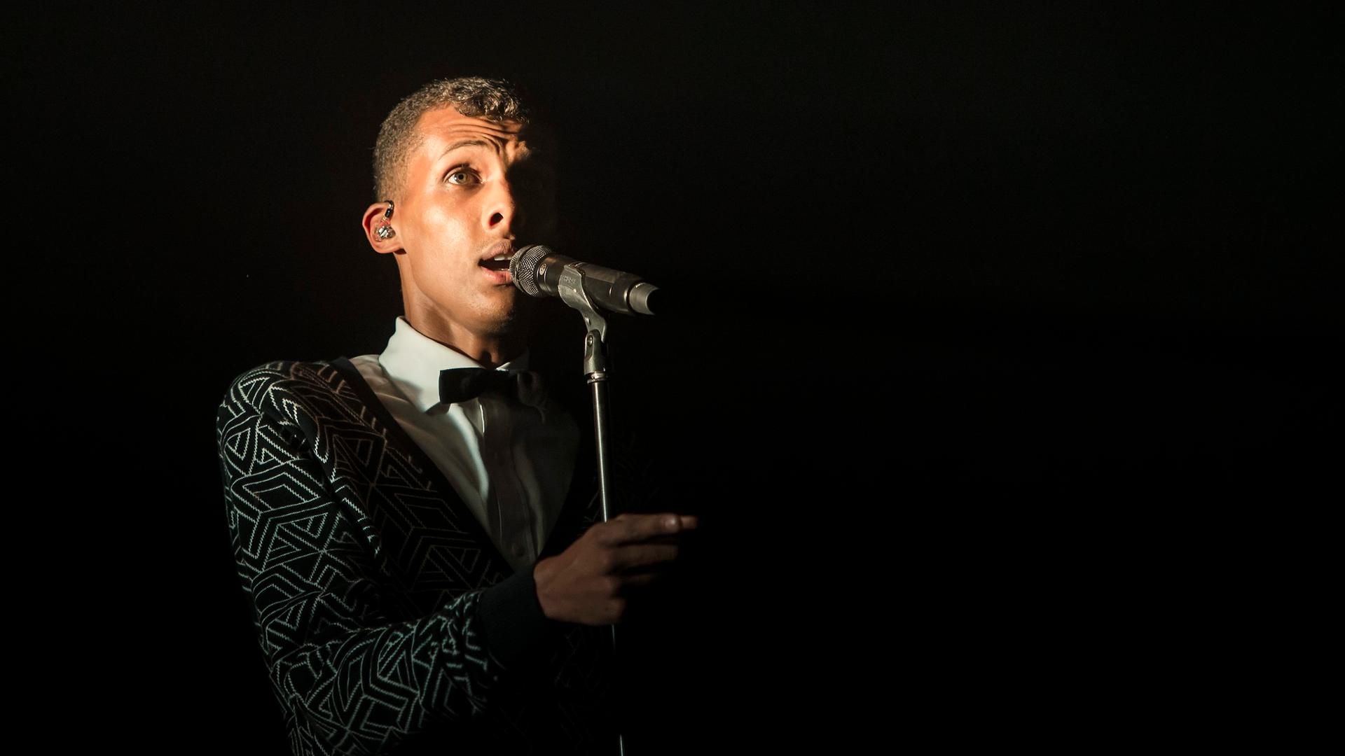 Der Sänger Stromae bei einem Konzert 2014: Er steht vor einem Mikro, der Hintergrund ist schwarz. Der Sänger Stromae bei einem Konzert 2014: Er steht vor einem Mikro, der Hintergrund ist schwarz.