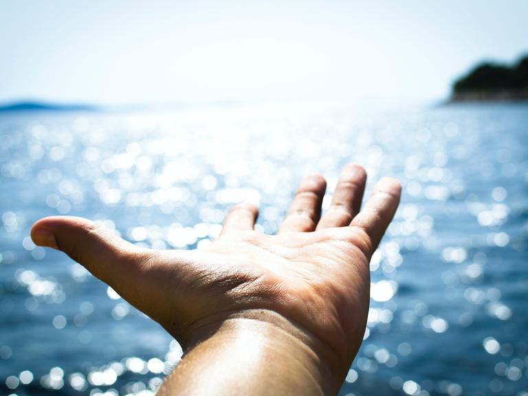 Hand zeigt mit geöffneter Handfläche Richtung Sonne und Meer. 