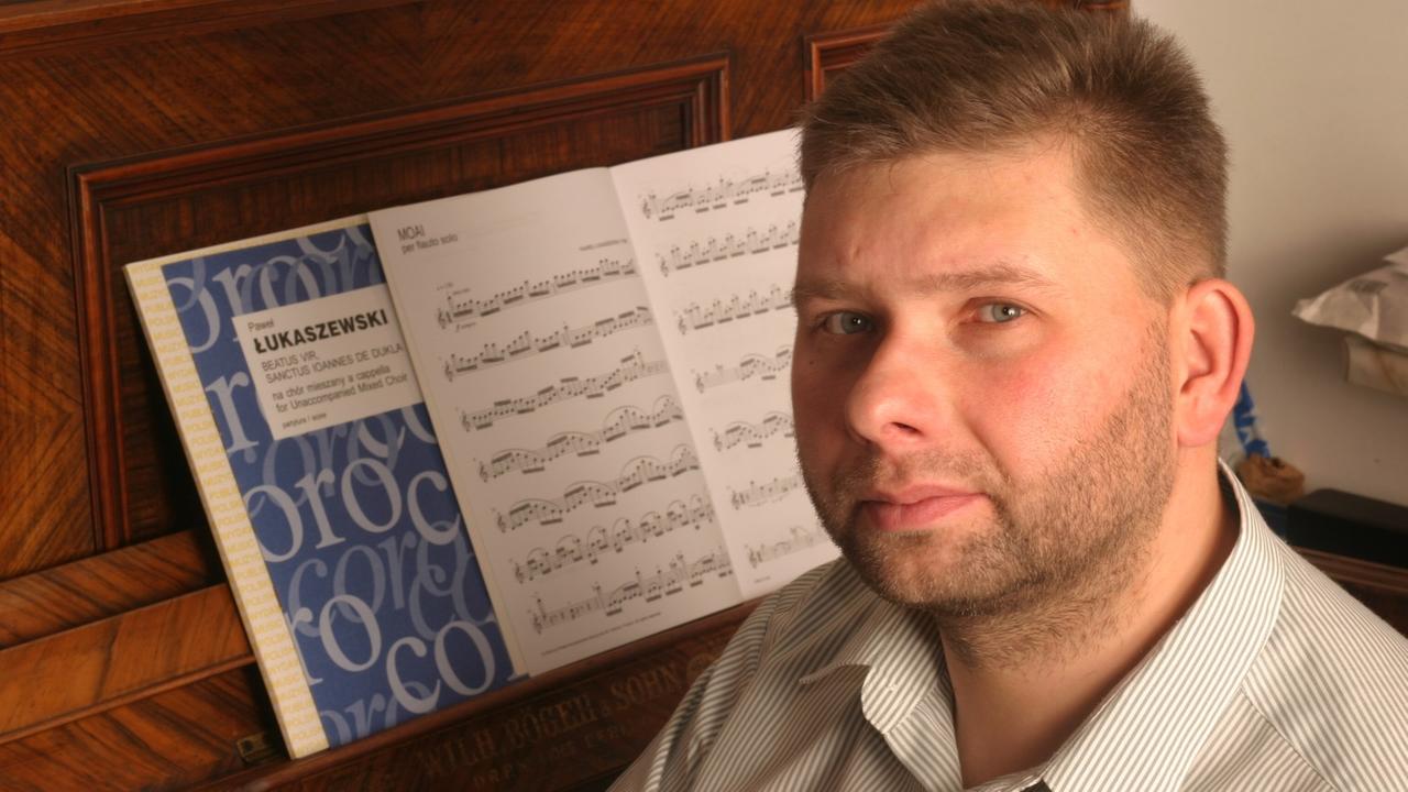 Ein Mann im gestreiften Hemd sitzt seitlich an einem Klavier mit aufgeblätterten Noten und schaut in die Kamera. Ein Mann im gestreiften Hemd sitzt seitlich an einem Klavier mit aufgeblätterten Noten und schaut in die Kamera.