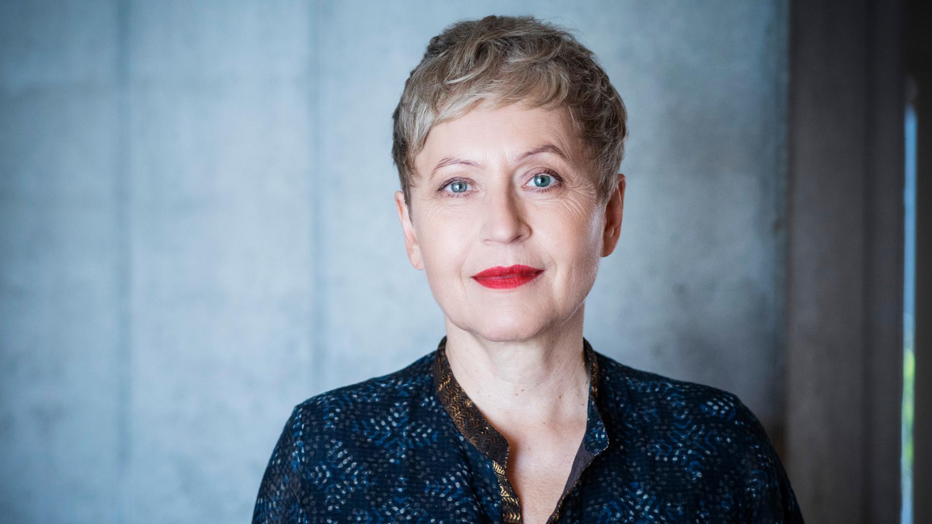 Ulrike Draesner schaut in die Kamera. Sie trägt ein dunkelblaue, gemusterte Bluse. Ihre Haare trägt sie kur, ihre Lippen hat sie rot geschminkt, sie hat blaue Augen. Ulrike Draesner schaut in die Kamera. Sie trägt ein dunkelblaue, gemusterte Bluse. Ihre Haare trägt sie kur, ihre Lippen hat sie rot geschminkt, sie hat blaue Augen.