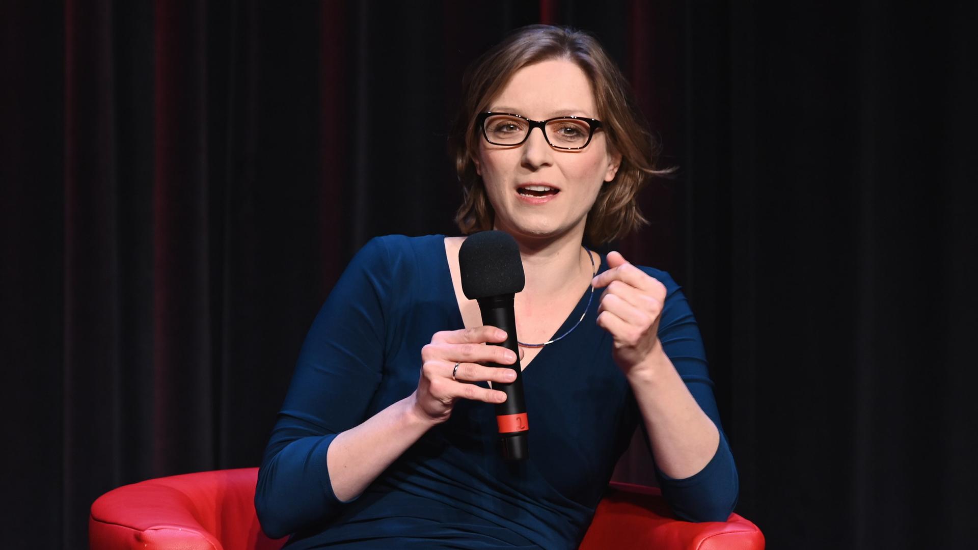 Die Philosophin Lisa Herzog sitzt auf einem Podium und spricht in ein Mikrofon. Die Philosophin Lisa Herzog sitzt auf einem Podium und spricht in ein Mikrofon.