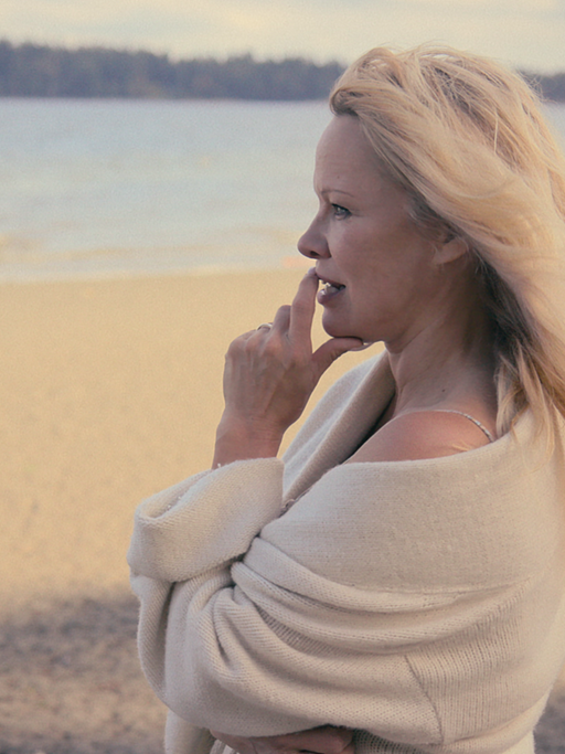 Die Schauspielerin Pamela Anderson steht an einem Sandstrand und schaut aufs Wasser. 