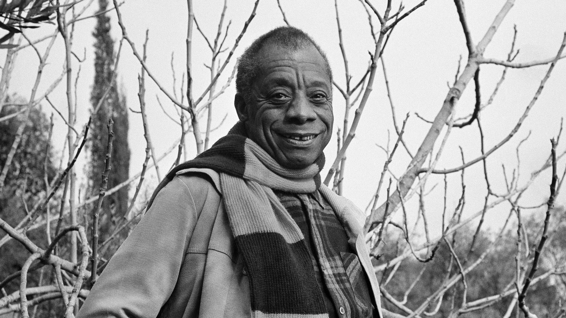 Der amerikanische Schriftsteller James Baldwin, fotografiert auf dem Grundstück seines Hauses an der französischen Riviera. Er trägt ein großkariertes Hemd, darüber eine offene Jacke und einen Schal.