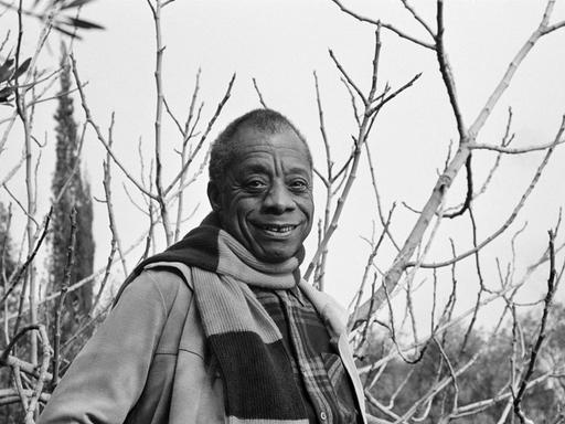 Der amerikanische Schriftsteller James Baldwin, fotografiert auf dem Grundstück seines Hauses an der französischen Riviera. Er trägt ein großkariertes Hemd, darüber eine offene Jacke und einen Schal.