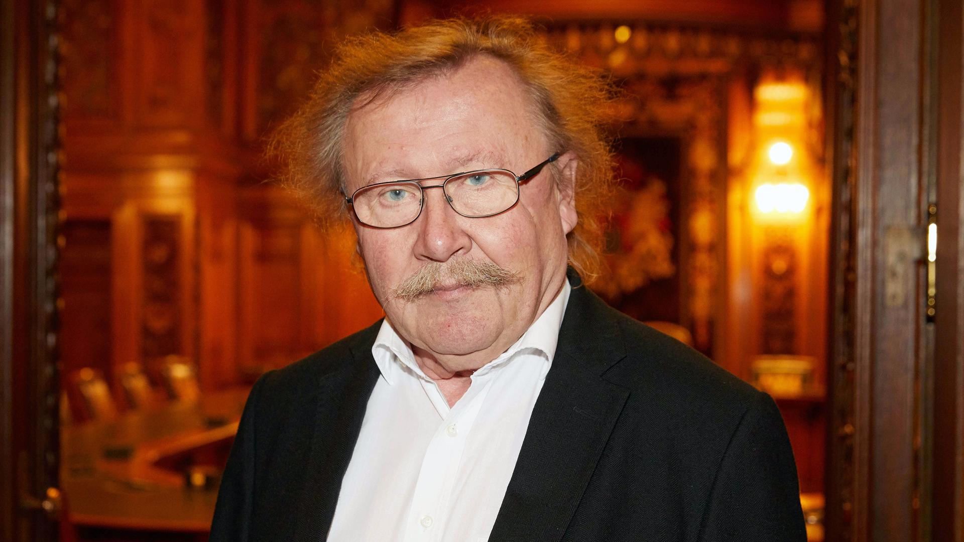 Porträt von Peter Sloterdijk, mit Brille und einem entschlossenen Blick in die Kamera. Im Hintergrund ist ein Sitzungssaal zu sehen, der in warmes Licht getaucht ist. Porträt von Peter Sloterdijk, mit Brille und einem entschlossenen Blick in die Kamera. Im Hintergrund ist ein Sitzungssaal zu sehen, der in warmes Licht getaucht ist.