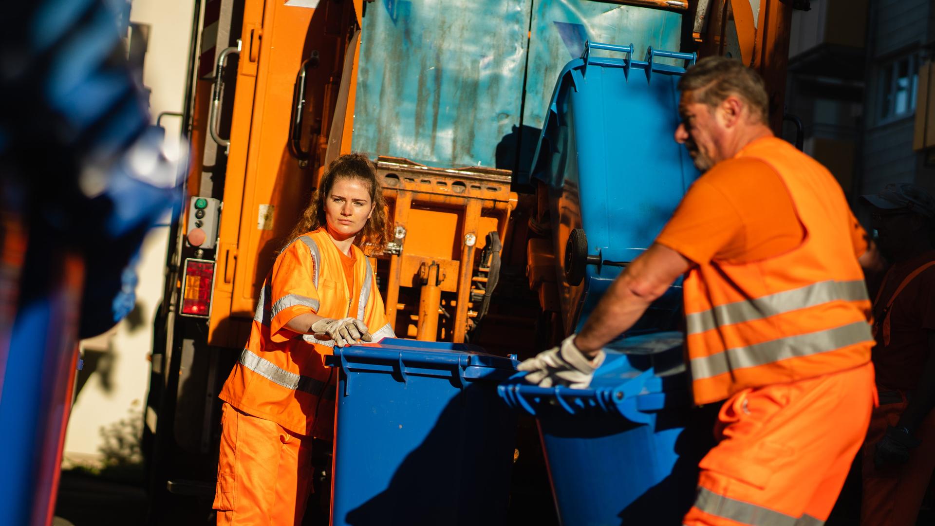 Das Foto zeigt 2 Müll-Werker bei der Arbeit. Zwei Müllwerker bei der Arbeit: Eine junge Frau und ein Mann schieben Papier-Mülltonnen zu einem Müll-Auto. 