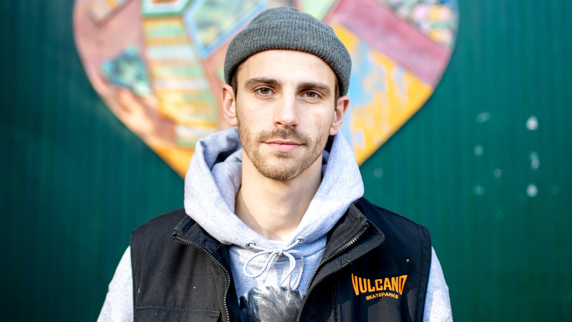 Fynn Kliemann, mit grauer Wollmütze, Kapuzenjacke und schwarzer Weste, steht vor einer grünen Wellblechwand mit einem stilisierten bunten Herz. Fynn Kliemann, mit grauer Wollmütze, Kapuzenjacke und schwarzer Weste, steht vor einer grünen Wellblechwand mit einem stilisierten bunten Herz.