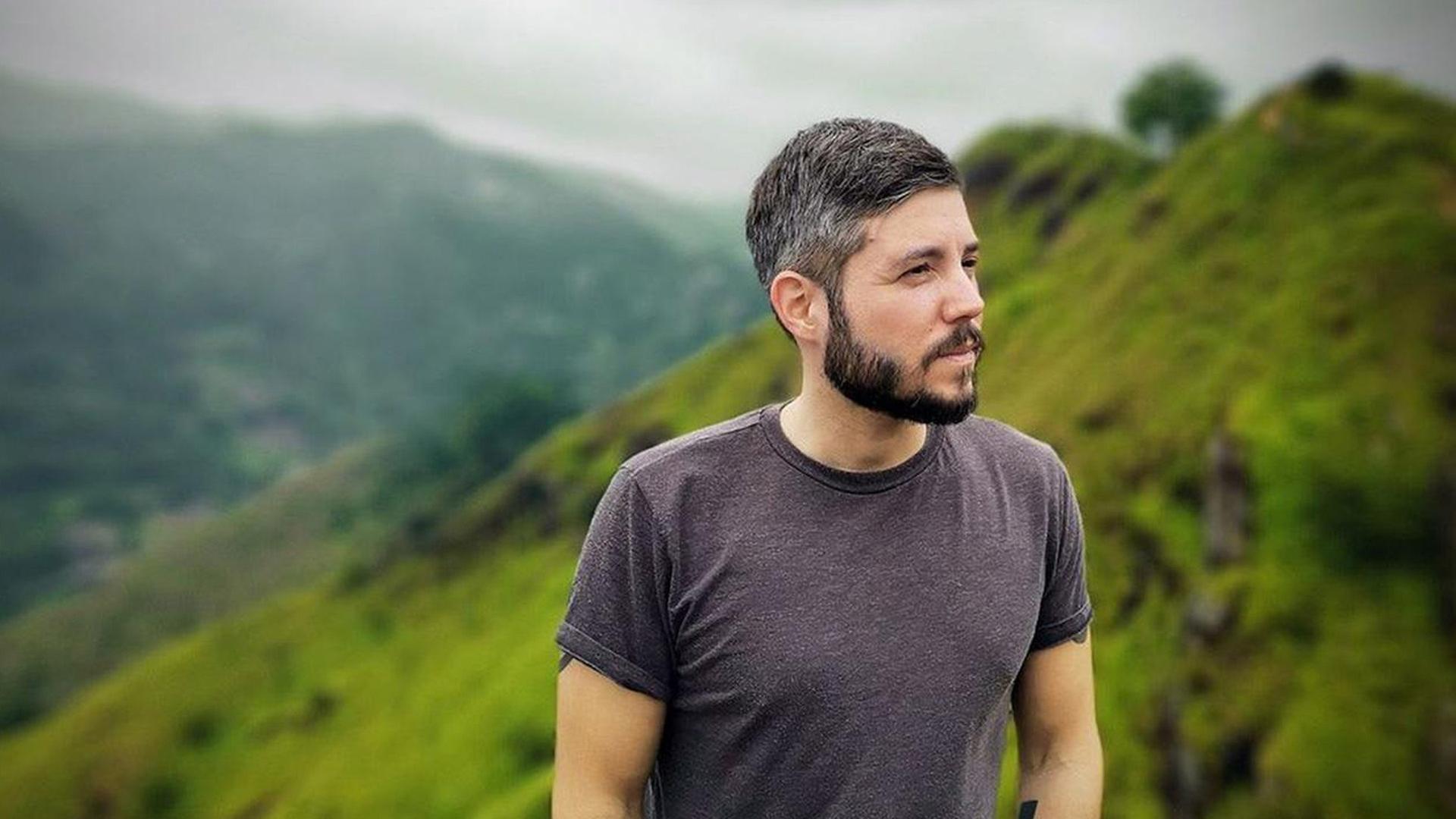 Ein junger Mann im Gebirge. Er hat ein T-Shirt an, trägt die haare kurz und hat einen Vollbart. Im Hintergrund sind saftige Weiden in einer Gebirgslandschaft zu sehen. Ein junger Mann im Gebirge. Er hat ein T-Shirt an, trägt die haare kurz und hat einen Vollbart. Im Hintergrund sind saftige Weiden in einer Gebirgslandschaft zu sehen.