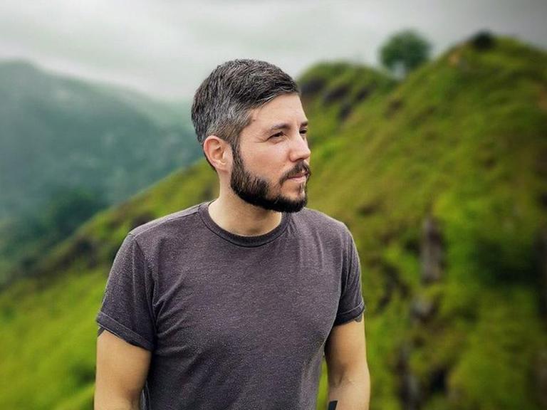 Ein junger Mann im Gebirge. Er hat ein T-Shirt an, trägt die haare kurz und hat einen Vollbart. Im Hintergrund sind saftige Weiden in einer Gebirgslandschaft zu sehen.