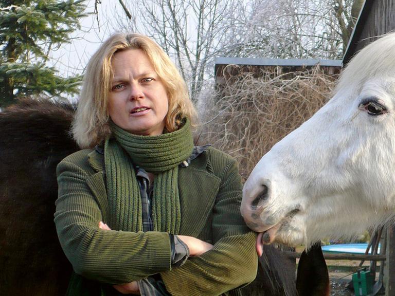 Die Schriftstellerin Karen Duve steht im Freien links neben einem Schimmel, dessen Kopf von rechts ins Bild ragt. Sie trägt eine hellgrüne Jacke und einen Wollschal in der gleichen Farbe.