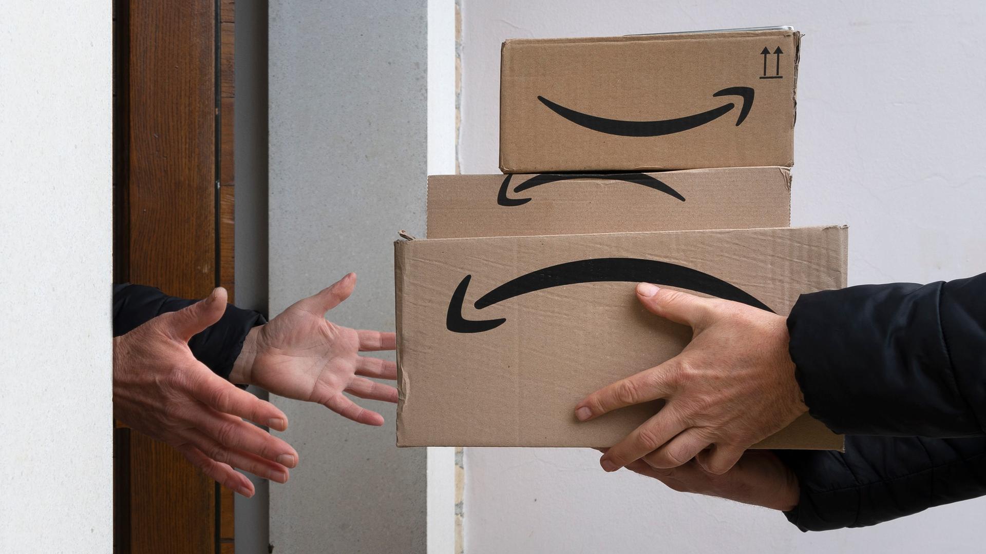 Drei Pakete mit dem Amazon Logo auf der Seite werden an einer Haustür überreicht. Drei Pakete mit dem Amazon Logo auf der Seite werden an einer Haustür überreicht.