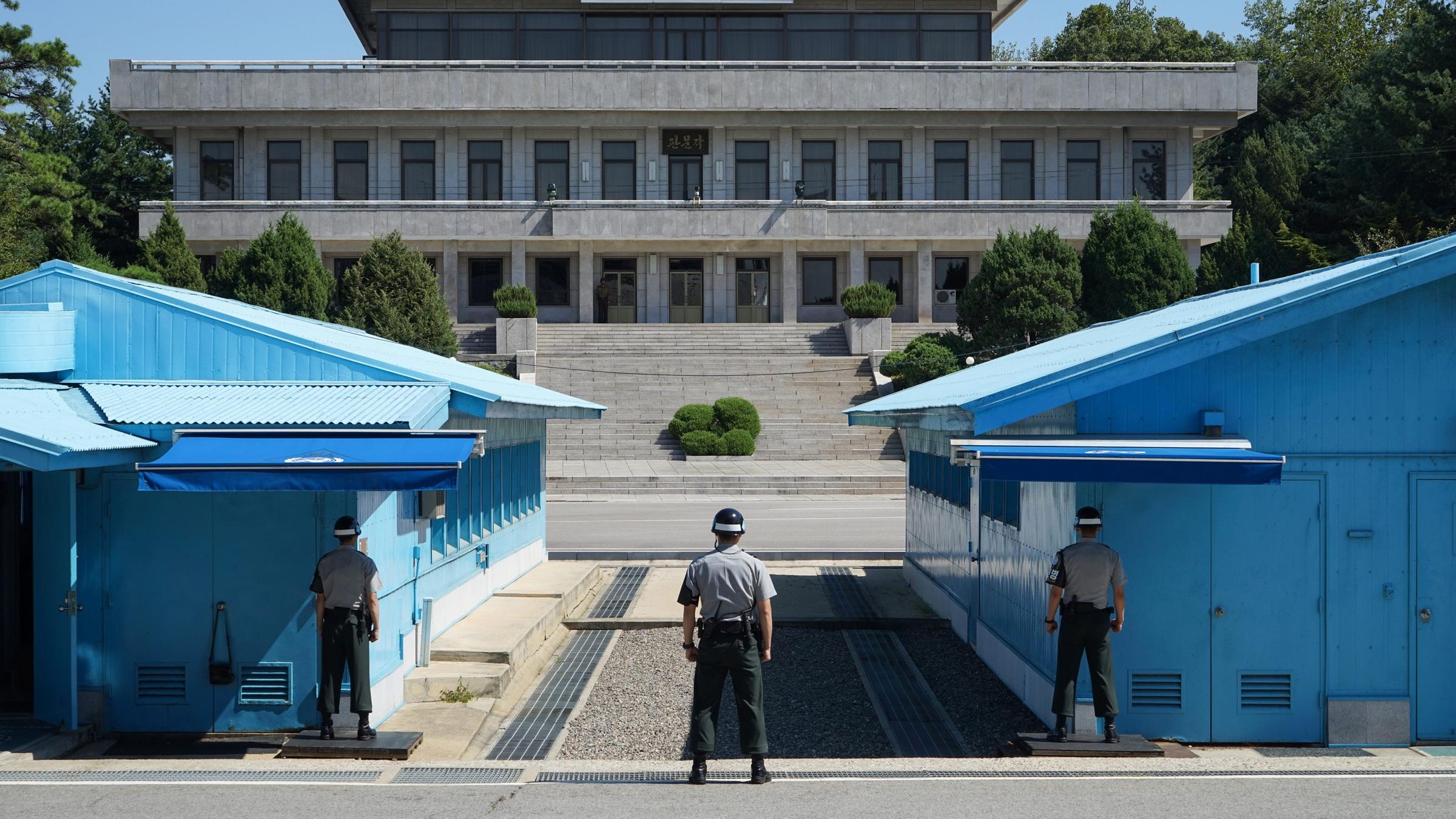Militärische Siedlung 'Panmunjeom' in der demilitarisierten Zone (DMZ) zwischen Nord- und Südkorea. Rechts und links sind blaue Baracken zu sehen. dazwischen verläuft die Grenze. im Vordergrund sind drei südkoreanische Grenzbeamten zu erkennen. Im Hintergrund sieht man ein Gebäude auf nordkoreanischer Seite.