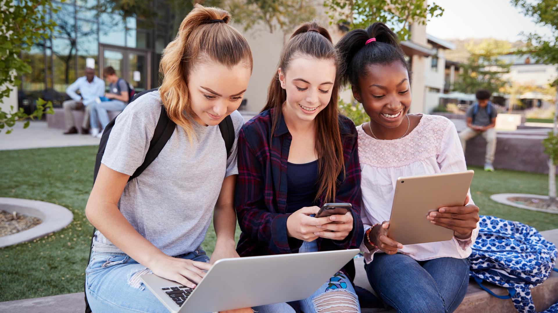 Drei Mädchen sitzen an einer Grünfläche nebeneinander, jede hat ein anderes Gerät in der Hand: Laptop, Smartphone, Tablet.