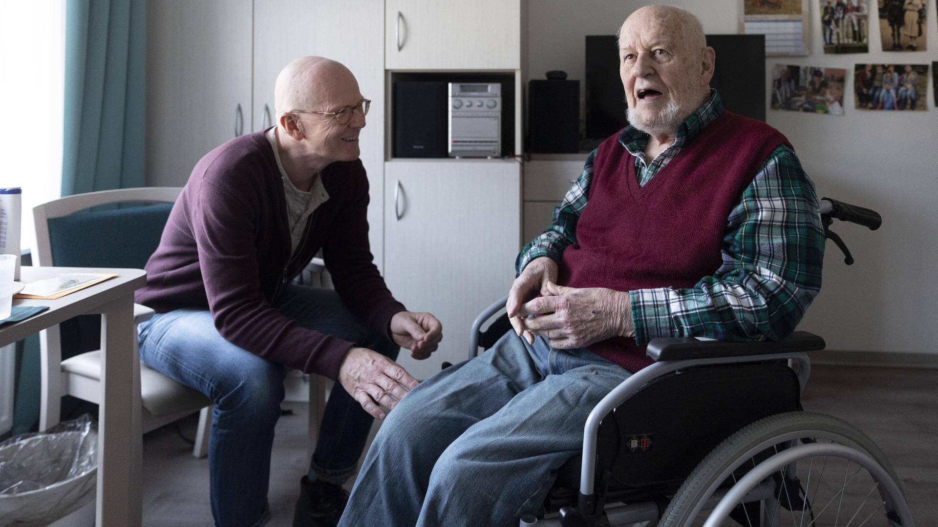Ein alter Mann sitzt im Pflegeheim im Rollstuhl, ein anderer Mann sitzt neben ihm. Beide sind im Gespräch.