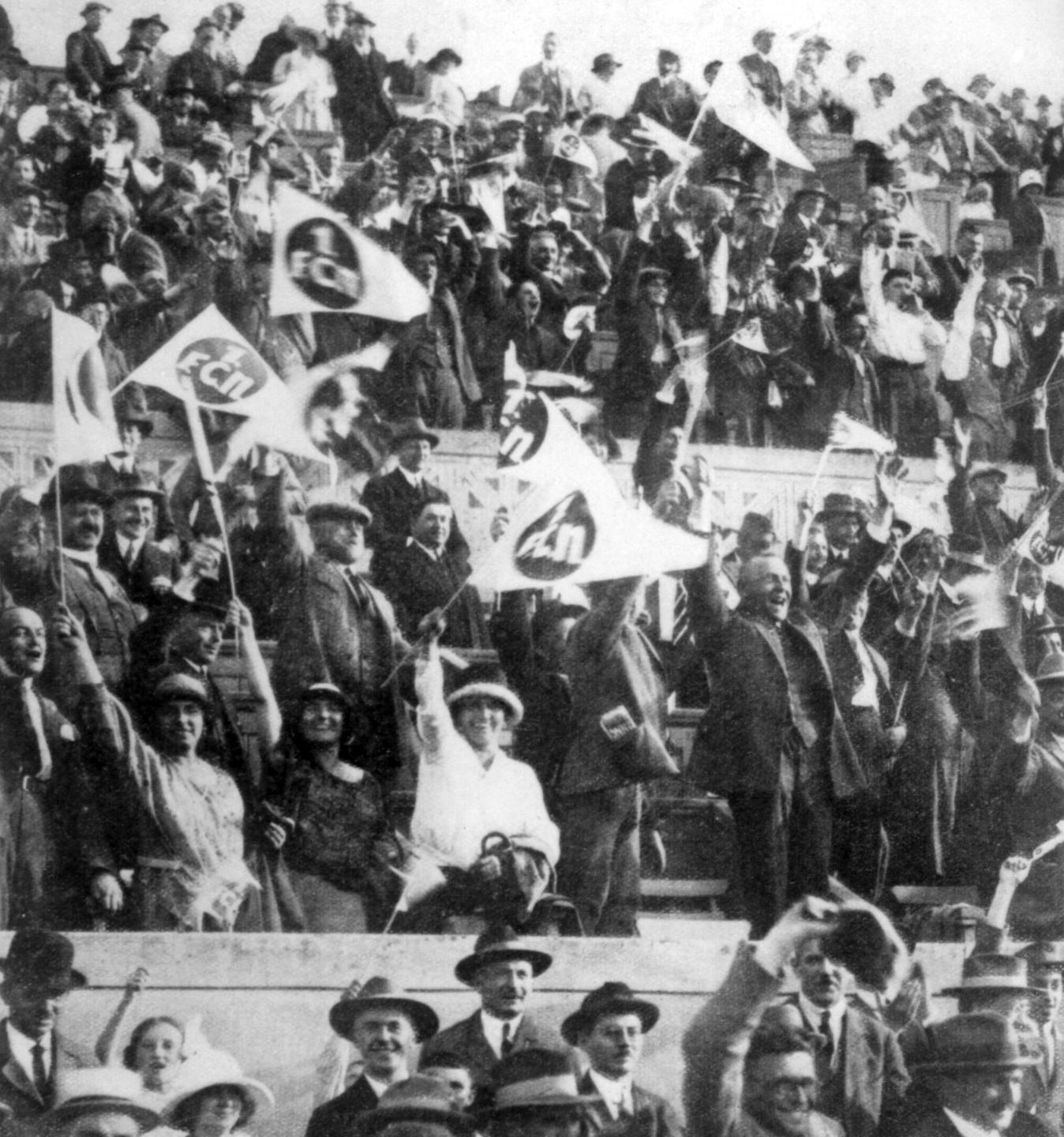 Schwarzweißfoto von einem historischen Fußballspiel. Auf einer Tribüne jubeln Fans ihrer Mannschaft zu. 