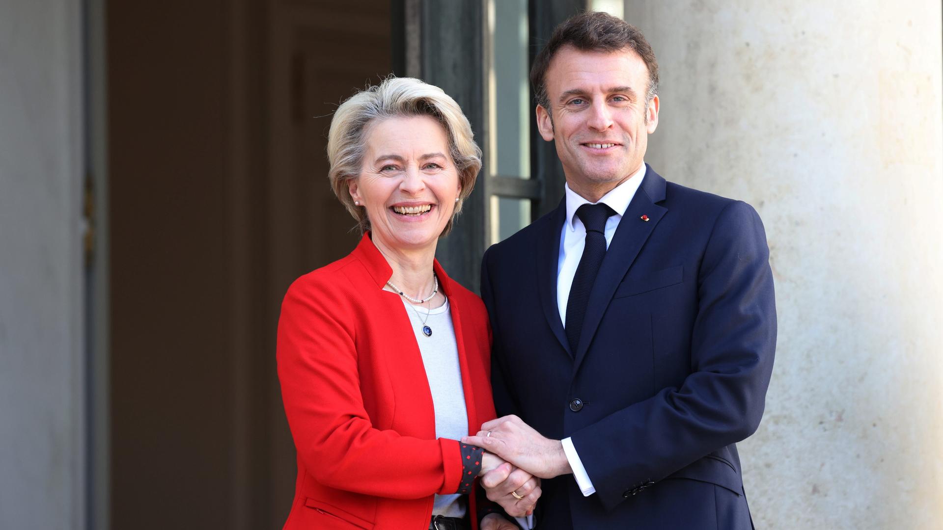 Ursula von der Leyen und Emmanuel Macron stehen Seite an Seite und lachen in die Kamera. Sie trägt einen roten Sack, er trägt einen blauen.