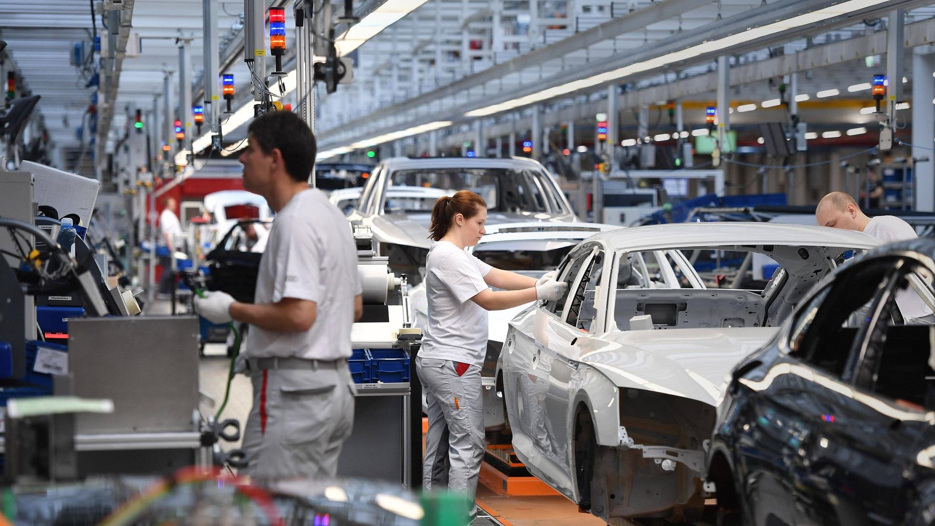 Eine Produktionsanlage von Audi. An den Karosserien arbeiten eine Frau und zwei Männer.