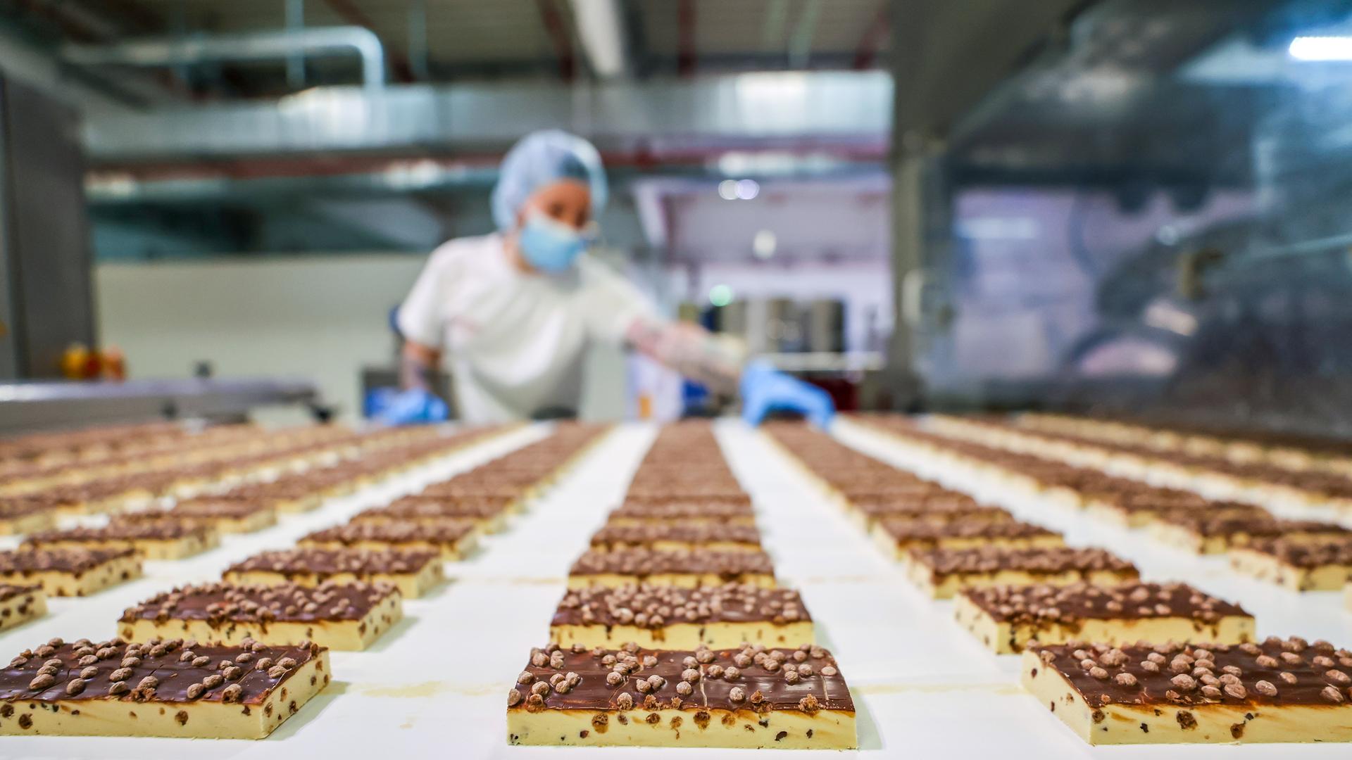 Ein Fließband mit vielen gleichmäßig aufgereiten Riegeln. Hinten im Bild ist ein Mitarbeiter mit Handschuhen zu erkennen, der die Produkte kontrolliert. Ein Fließband mit vielen gleichmäßig aufgereiten Riegeln. Hinten im Bild ist ein Mitarbeiter mit Handschuhen zu erkennen, der die Produkte kontrolliert.