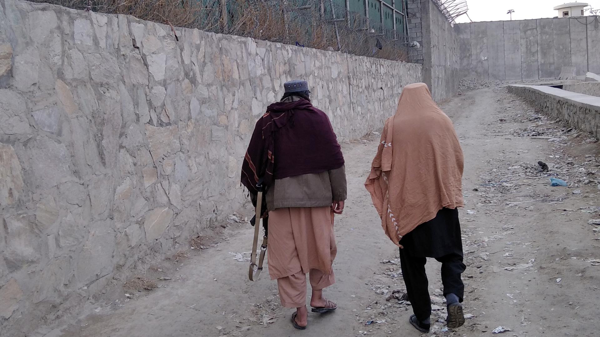 Zwei Männer laufen an einer hohen Mauer mit Stacheldraht, einer trägt ein Gewehr unter dem Arm, Afghanistan 2022. Zwei Männer laufen an einer hohen Mauer mit Stacheldraht, einer trägt ein Gewehr unter dem Arm, Afghanistan 2022.