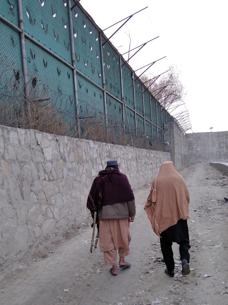 Zwei Männer laufen an einer hohen Mauer mit Stacheldraht, einer trägt ein Gewehr unter dem Arm, Afghanistan 2022.